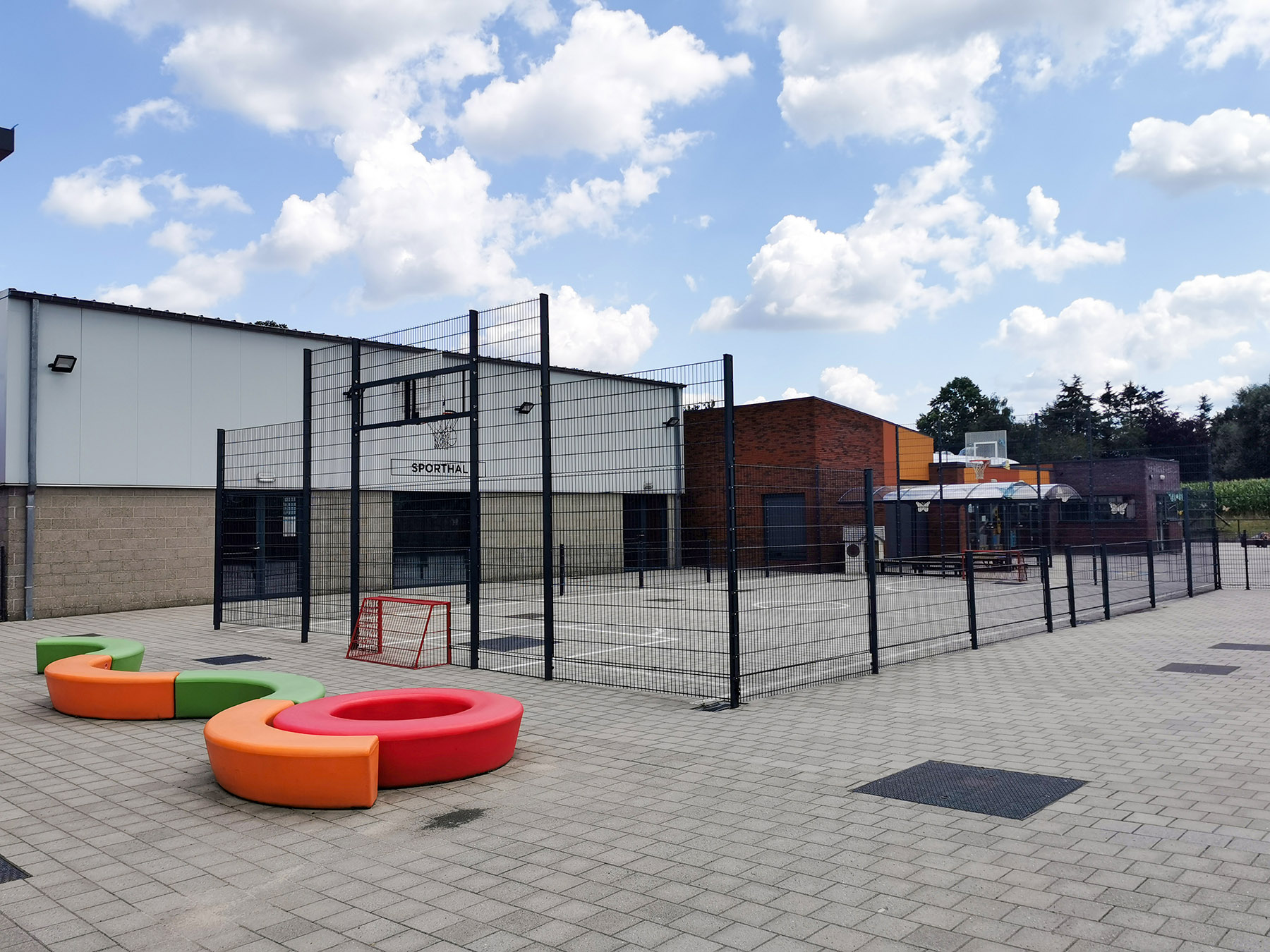 Sportkooi met basketbalring en zitbanken op de speelplaats, naast de sporthal